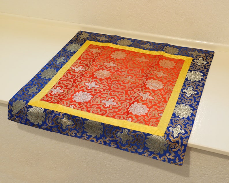 Puede incluir: Un coj&iacute;n de meditaci&oacute;n rojo y dorado con un borde de patr&oacute;n floral azul y plateado. El coj&iacute;n est&aacute; hecho de tela y tiene una forma cuadrada.
