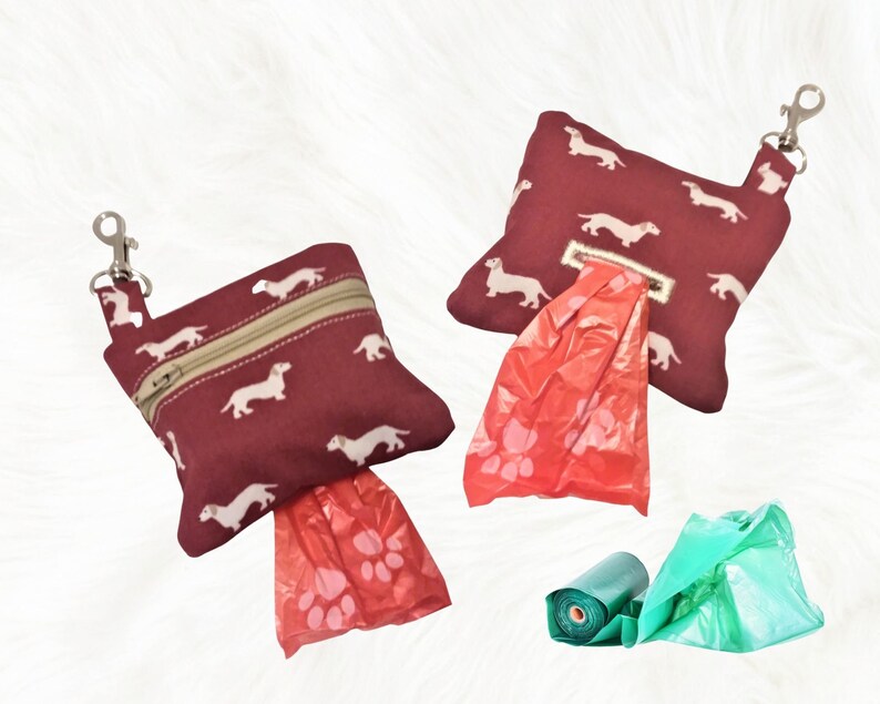 May include: Two red dog poop bag dispensers with dachshund prints and a silver clip. One dispenser has a red bag with paw prints and the other has a green bag.