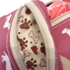 May include: A pink zippered pouch with a white dog print and a beige zipper. The pouch has a beige lining with a paw print and bone pattern. The pouch is open and a pink bag is partially visible inside.