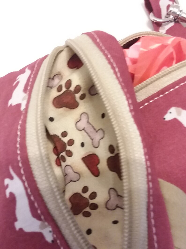 May include: A pink zippered pouch with a white dog print and a beige zipper. The pouch has a beige lining with a paw print and bone pattern. The pouch is open and a pink bag is partially visible inside.