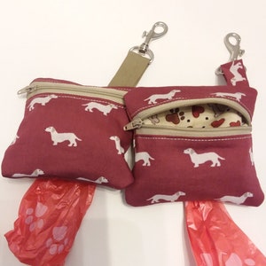 May include: Two burgundy fabric dog poop bag holders with dachshund print and beige trim. Each holder has a zipper closure and a silver clip. The holders are attached to a beige strap with a silver clip. Two red plastic poop bags are included.