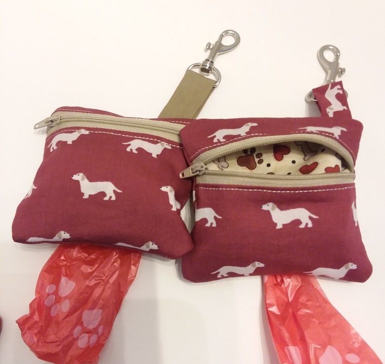 May include: Two burgundy fabric dog poop bag holders with dachshund print and beige trim. Each holder has a zipper closure and a silver clip. The holders are attached to a beige strap with a silver clip. Two red plastic poop bags are included.