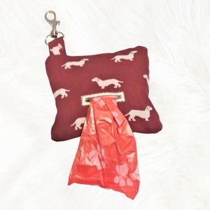 May include: A burgundy dog poop bag dispenser with a white dachshund print and a silver clip. The dispenser has a red plastic bag with paw prints inside.