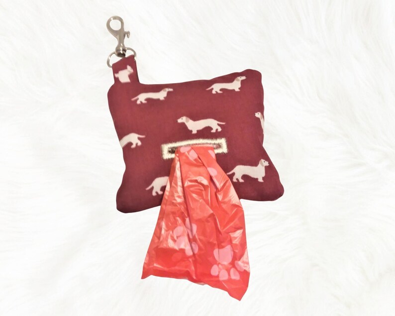 May include: A burgundy dog poop bag dispenser with a white dachshund print and a silver clip. The dispenser has a red plastic bag with paw prints inside.