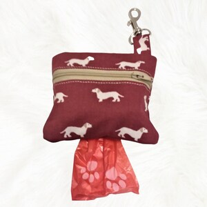May include: A burgundy zippered pouch with a white dachshund print and a silver clip. The pouch is filled with red plastic dog waste bags with paw prints.