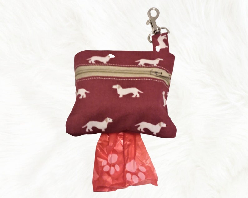 May include: A burgundy zippered pouch with a white dachshund print and a silver clip. The pouch is filled with red plastic dog waste bags with paw prints.