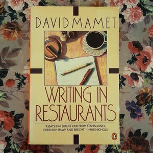 May include: A paperback book titled Writing in Restaurants by David Mamet. The cover features an illustration of a desk with a cup of coffee, a pencil, a pen, and a cigar. The text includes a quote by Mike Nichols and a small penguin logo.