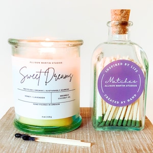 May include: A lit candle in a glass jar with a label that reads "Sweet Dreams." Next to it is a glass bottle filled with matches, and a burnt match on a wooden surface. The candle is 4 oz / 113 g.