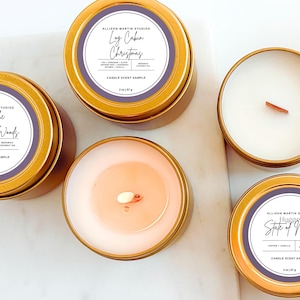 Op de afbeelding: Verschillende goudkleurige, ronde kaarsenblikken met witte etiketten en tekst. De etiketten luiden "Oh to be Lost in the Woods", "Log Cabin Christmas" en "Hygge State of Mind". De kaarsen zijn elk 85 g.