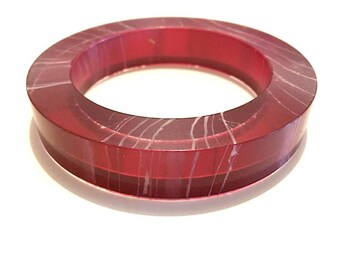 May include: A thick, circular bangle bracelet in a deep red hue. The bracelet features subtle, white, linear patterns throughout its surface. The design is simple and elegant, suitable for everyday wear. The bangle is approximately 1 inch wide.