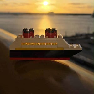 May include: A toy cruise ship constructed from colorful building blocks. The ship features a white hull with black and yellow accents, and red and black smokestacks. The background shows a sunset over the water.