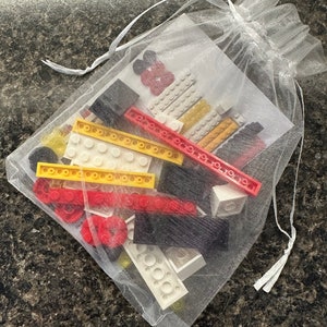 May include: A white mesh drawstring bag filled with colorful toy building blocks. The blocks are red, yellow, black, and white. The bag is tied with a white ribbon.