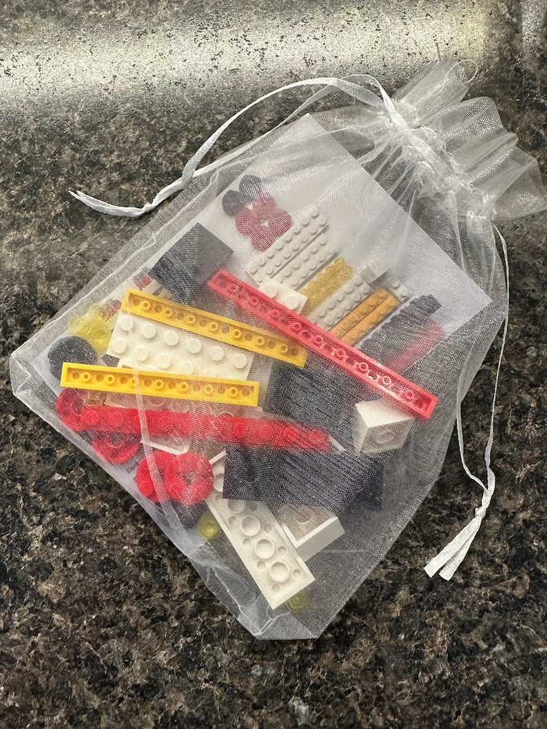 May include: A white mesh drawstring bag filled with colorful toy building blocks. The blocks are red, yellow, black, and white. The bag is tied with a white ribbon.