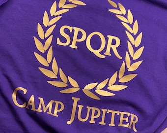 Puede incluir: Camiseta morada con un diseño de corona de laurel dorada. El texto "SPQR" está dentro de la corona, y "CAMP JUPITER" debajo. La camiseta está anudada en la parte inferior.