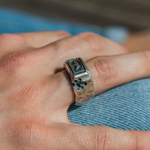 May include: A silver ring with a textured, hammered finish. The ring features a rectangular design with a floral motif.