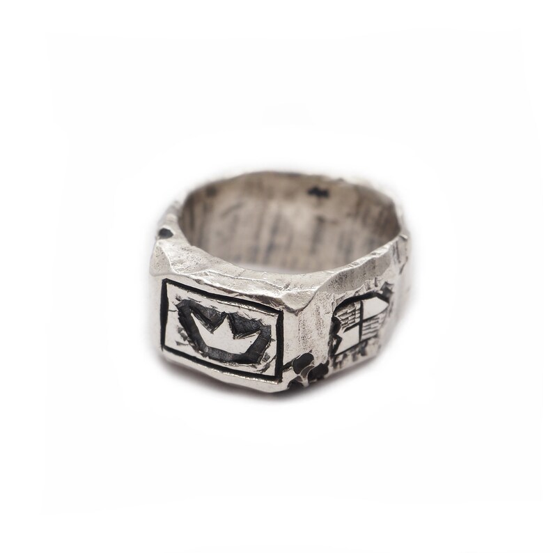 May include: A silver ring with a textured surface and a crown design in the center. The ring has a rectangular shape and is decorated with other designs.