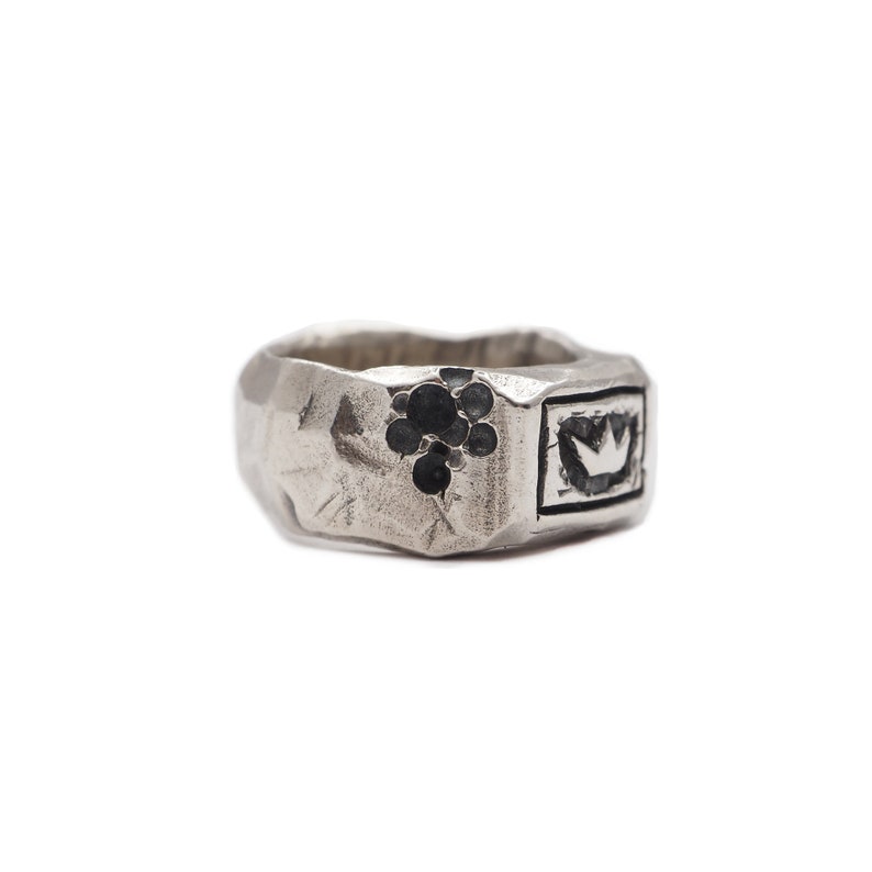 May include: A silver ring with a textured band and a black enamel design of a crown and grapes.