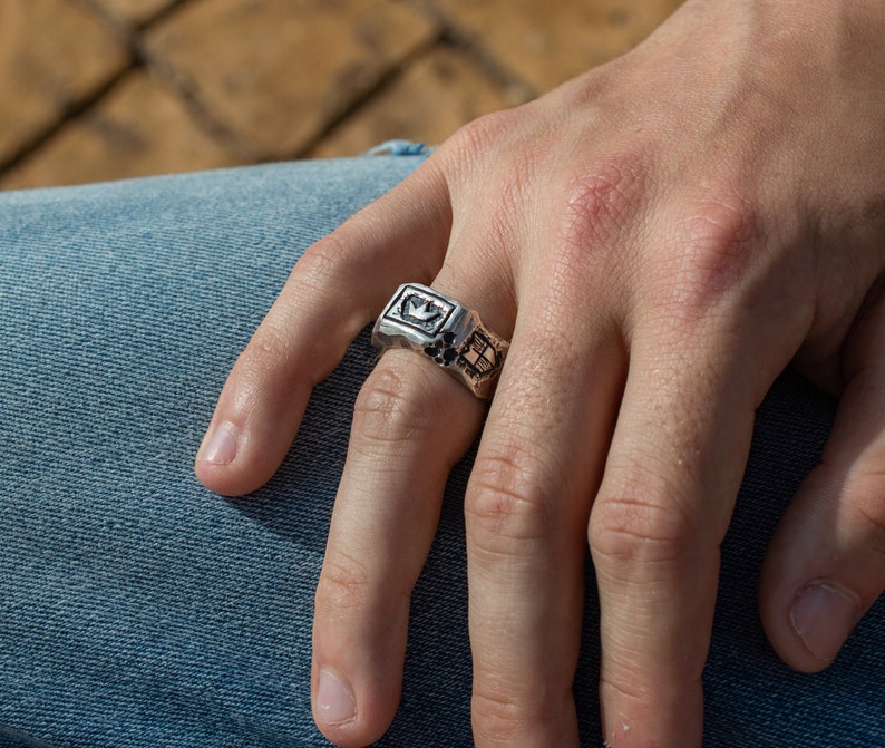 May include: A silver ring with a crown design and intricate details. The ring is worn on the index finger of a hand.