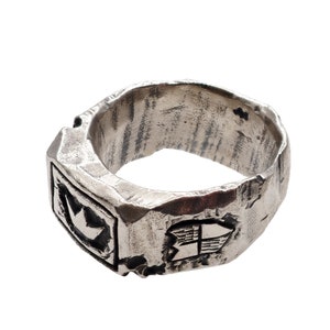 May include: A silver ring with a textured surface and two engraved designs. One design features a crown and the other a shield with a cross.