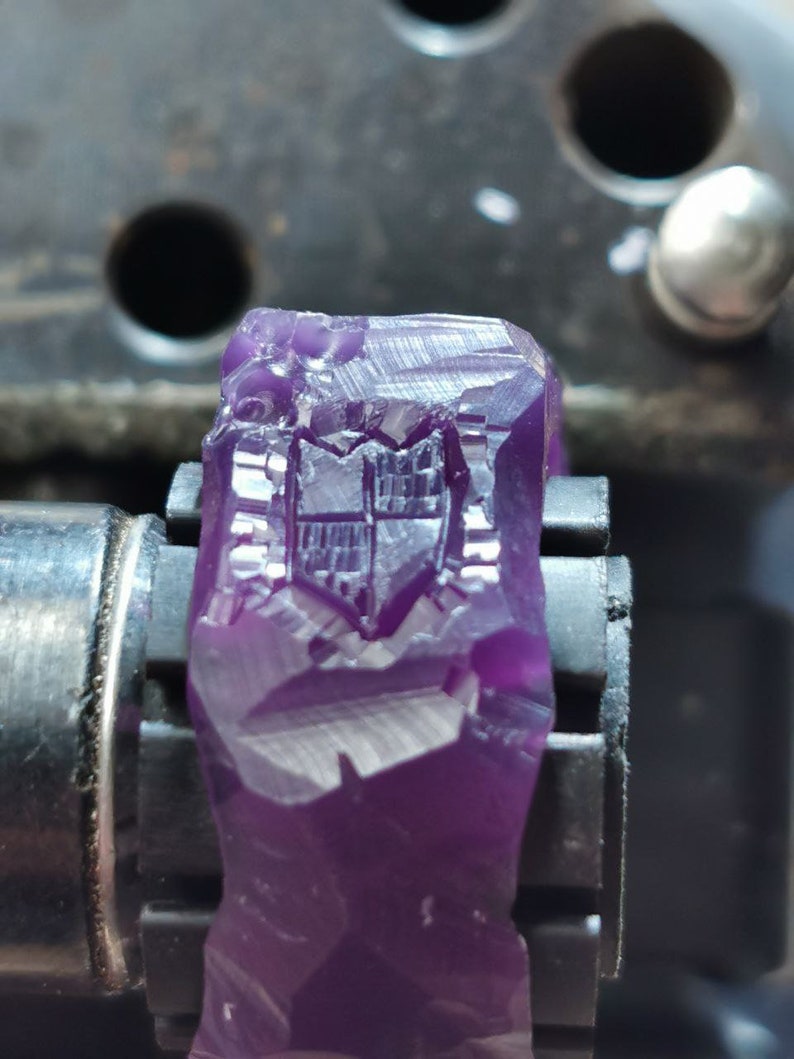 May include: A close-up of a purple wax ring with a detailed design carved into the surface. The ring is being held in a metal clamp.