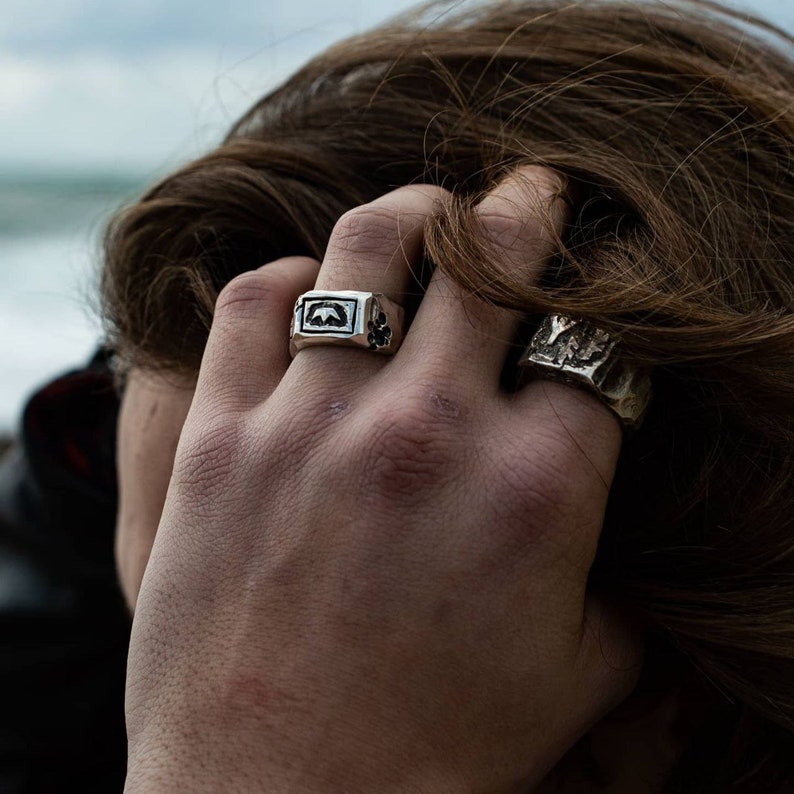 May include: A person's hand wearing two silver rings. The ring on the index finger has a black and white design, while the ring on the pinky finger has a more intricate design.