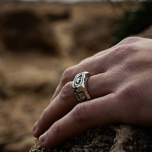 May include: A silver ring with a detailed design featuring a crescent moon and other geometric shapes. The ring is worn on a person's finger.