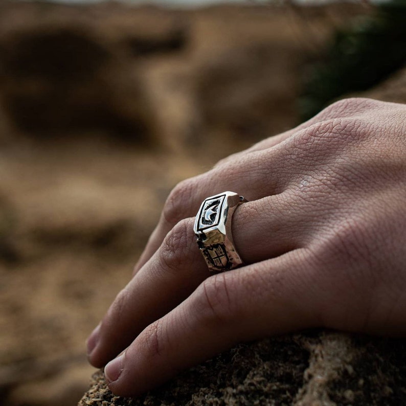 May include: A silver ring with a detailed design featuring a crescent moon and other geometric shapes. The ring is worn on a person's finger.