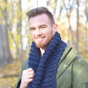 Op de afbeelding: Een man draagt een donkerblauwe gebreide sjaal en een groene jas. De sjaal is dik en grof gebreid, en de jas heeft een rits en zakken.