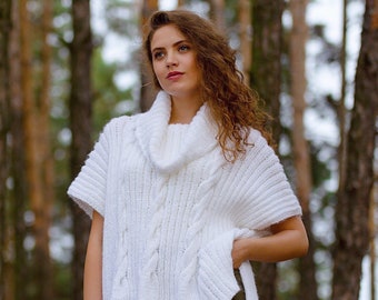 May include: A white cable-knit sweater with a cowl neck and short sleeves. The sweater features a textured design and a loose fit. The image is set in a forest, with the text "Designed by COZYYARNstudio" at the bottom.