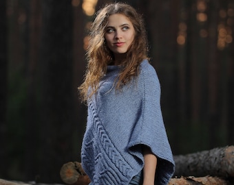 May include: A woman wearing a blue knitted poncho with a cable knit pattern, standing in a forest setting. The poncho has a loose fit and a long, flowing design. The woman is looking off to the side, and her hair is long and flowing.