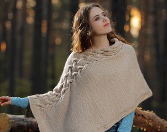 May include: A woman wearing a beige knitted poncho with a cable knit design. She is sitting on a pile of logs in a woodland setting.