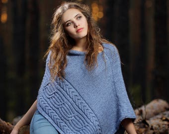 May include: A woman wearing a blue knitted poncho with a cable knit design. She is sitting on a pile of logs in a woodland setting.
