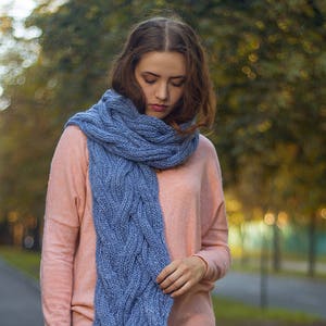 May include: A woman wearing a light pink sweater with a blue knitted scarf. The scarf has a cable knit pattern.