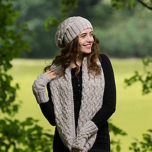 May include: A light gray knit hat, scarf, and fingerless gloves set. The scarf and gloves have a cable knit design. The set is designed by COZYYARNStudio. The person is wearing a black long-sleeve shirt and black pants.