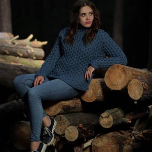 May include: A woman wearing a blue knitted sweater and jeans sits on a pile of logs in a forest setting.