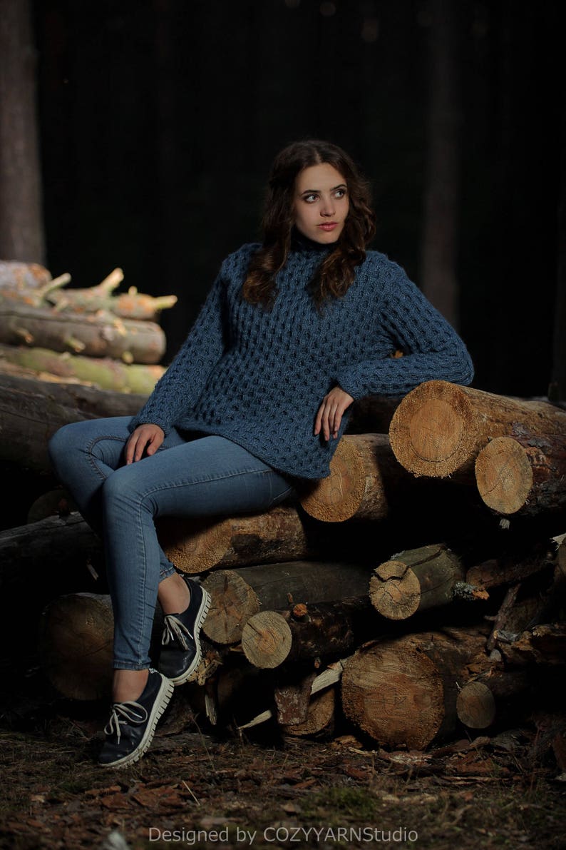 May include: A woman wearing a blue knitted sweater and jeans sits on a pile of logs in a forest setting.