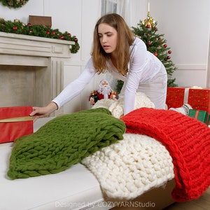 Puede incluir: Una mujer con una camisa blanca est&aacute; alcanzando una caja de regalo roja envuelta con una cinta dorada. La caja de regalo est&aacute; en un sof&aacute; blanco con una manta de punto verde, blanca y roja.