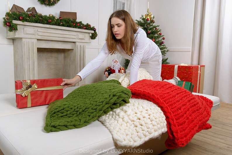 Puede incluir: Una mujer con una camisa blanca est&aacute; alcanzando una caja de regalo roja envuelta con una cinta dorada. La caja de regalo est&aacute; en un sof&aacute; blanco con una manta de punto verde, blanca y roja.