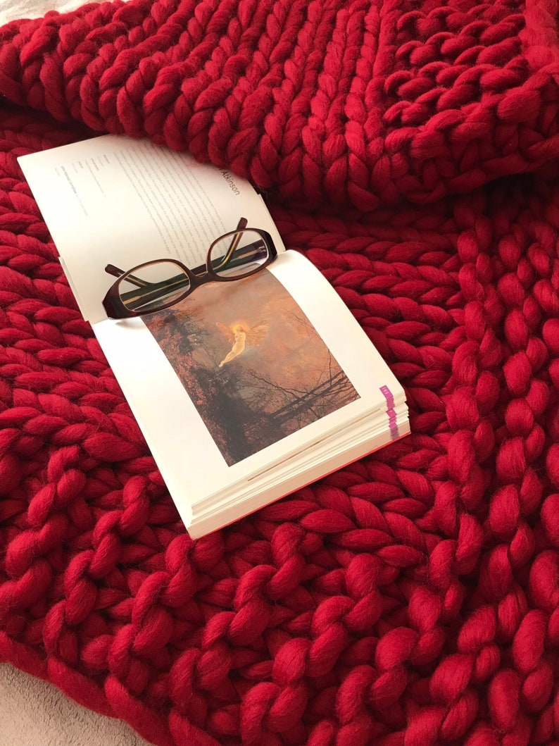 Puede incluir: Primer plano de una manta de punto gruesa de color rojo con un libro y unas gafas encima. La manta est&aacute; hecha de hilo grueso y tiene una textura suave y acogedora.