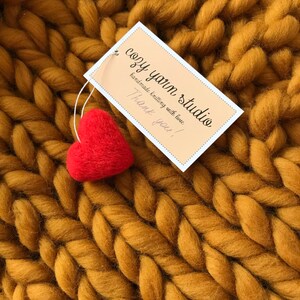 Puede incluir: Primer plano de una manta de punto grueso de color amarillo mostaza. Un peque&ntilde;o coraz&oacute;n de fieltro rojo se coloca encima de la manta. Una etiqueta blanca con el texto "cozy yarn studio handmade knitting with love Thank you!" est&aacute; unida a la manta.