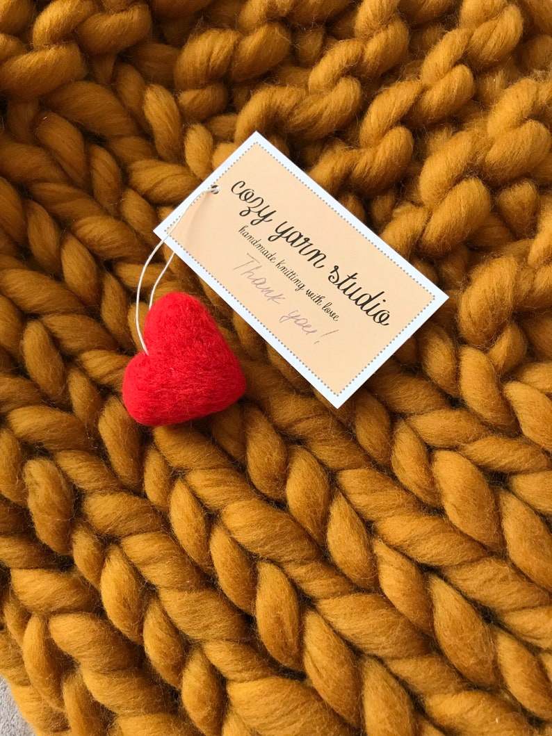 Puede incluir: Primer plano de una manta de punto grueso de color amarillo mostaza. Un peque&ntilde;o coraz&oacute;n de fieltro rojo se coloca encima de la manta. Una etiqueta blanca con el texto "cozy yarn studio handmade knitting with love Thank you!" est&aacute; unida a la manta.