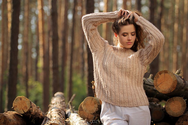 May include: A woman wearing a light brown knitted sweater with a cable knit pattern stands in a forest. She is holding her hair up with her hands. The background is a blurred image of trees.