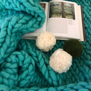 Puede incluir: Una manta de punto grueso de color turquesa con pompones blancos y verdes. Un libro abierto con una pintura de un paisaje est&aacute; debajo de la manta.