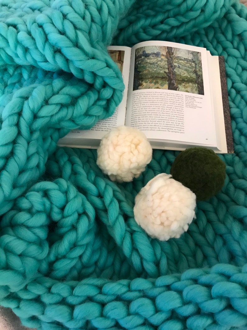 Puede incluir: Una manta de punto grueso de color turquesa con pompones blancos y verdes. Un libro abierto con una pintura de un paisaje est&aacute; debajo de la manta.