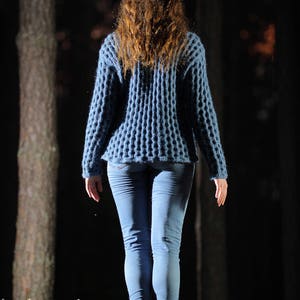 May include: A woman wearing a blue knitted sweater and blue jeans walks through a dark forest.