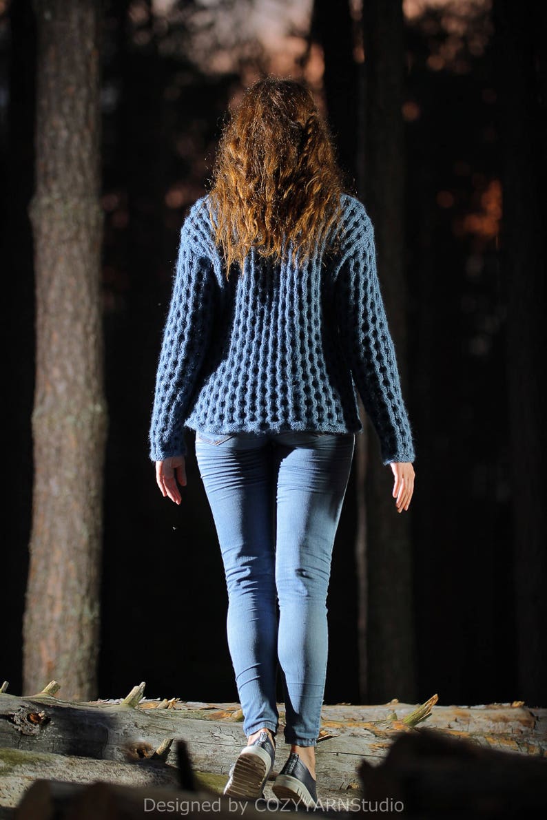 May include: A woman wearing a blue knitted sweater and blue jeans walks through a dark forest.