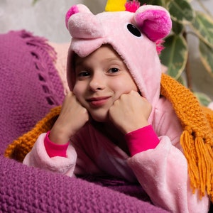 May include: A young person wearing a pink unicorn onesie with a yellow horn and pink ears. They are lying on a purple knitted blanket and looking at the camera with a smile.