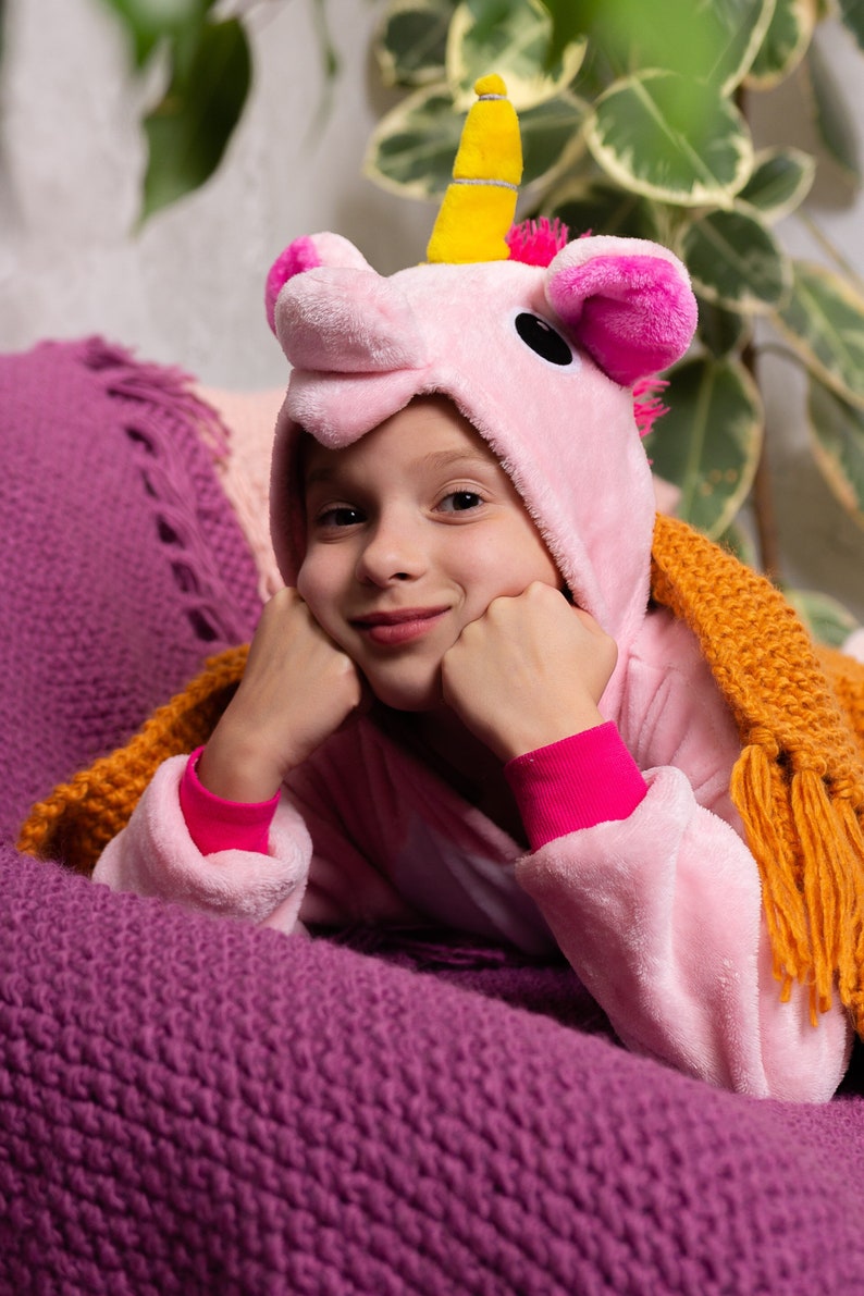 May include: A young person wearing a pink unicorn onesie with a yellow horn and pink ears. They are lying on a purple knitted blanket and looking at the camera with a smile.