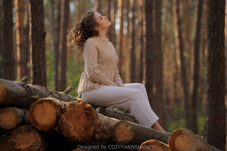 May include: A woman wearing a beige knitted sweater and white pants sits on a pile of logs in a forest. The sun shines through the trees, creating a warm glow.