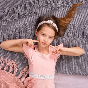 May include: A young girl with long brown hair wearing a pink dress with a white lace trim lies on a gray and pink blanket. She is looking at the camera and has her fingers on her cheeks.